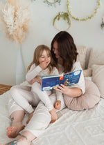 A mom and her daughter reading a book in bed while wearing matching neutral PJ's from Milana Label.