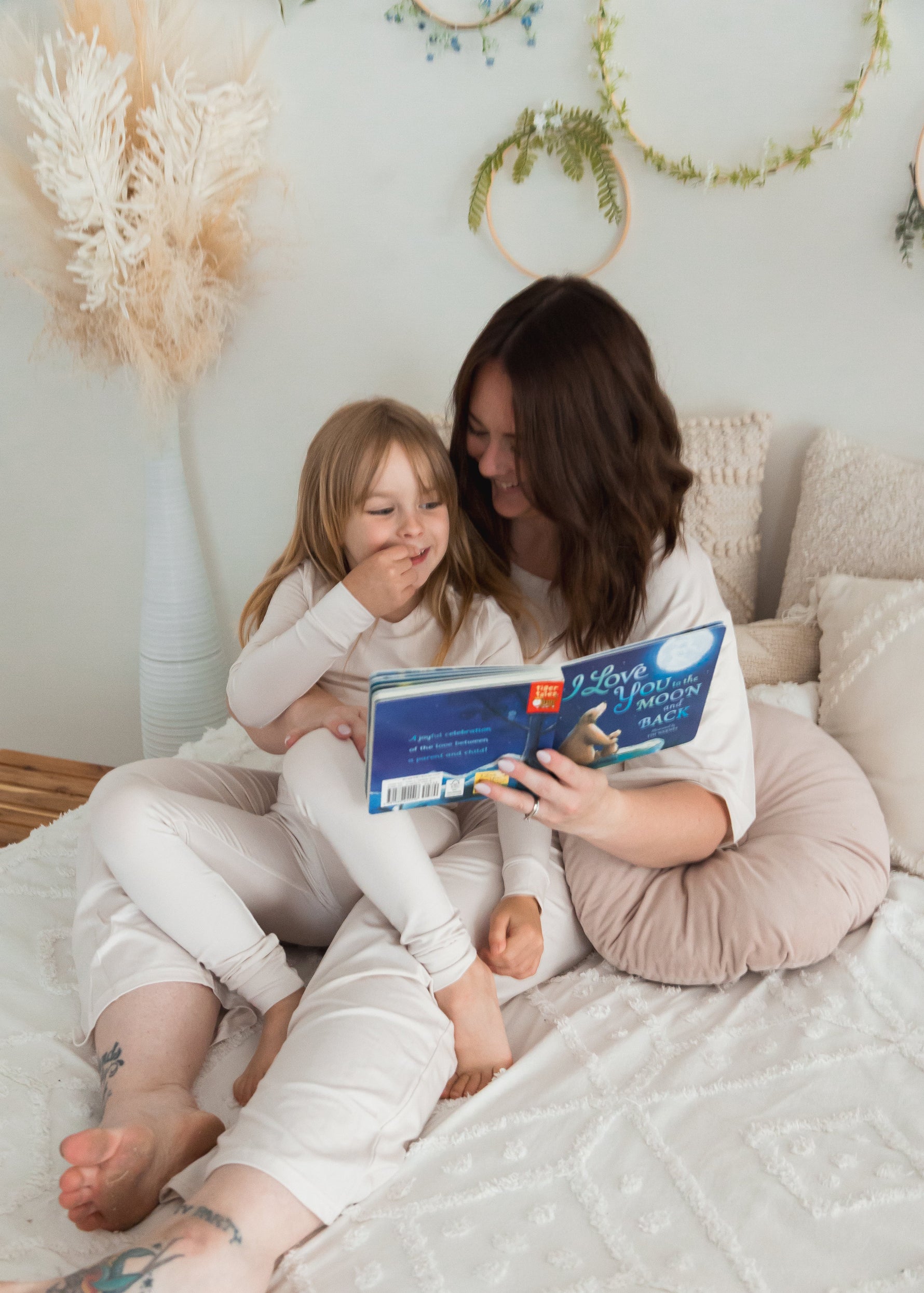 A mom and her daughter reading a book in bed while wearing matching neutral PJ's from Milana Label.