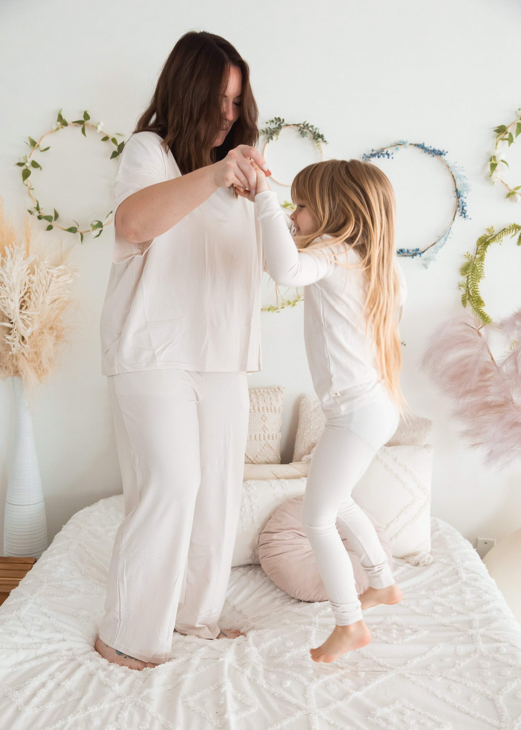 Vertical shot of a mother and her young daughter holding hands and jumping playfully on a white textured bedspread, both wearing matching Milana Label mommy and me loungewear sets in Birch.