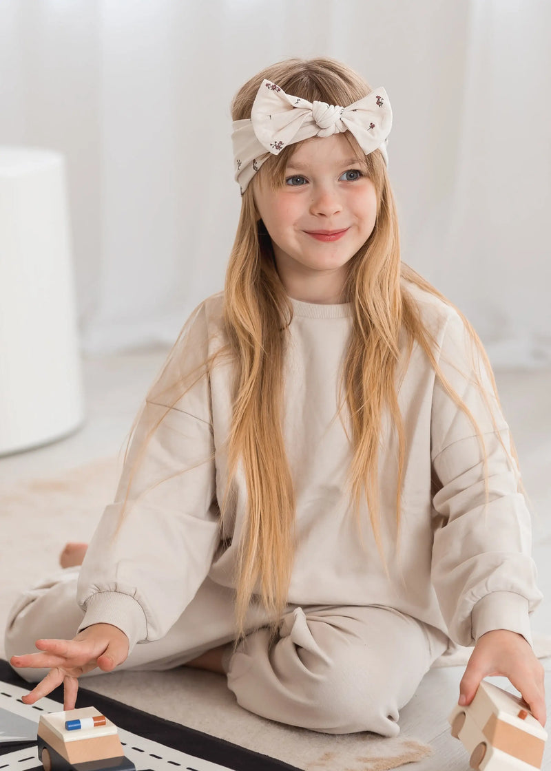 A young girl with long blonde hair wearing an organic cotton, oat-colored jogger set. She sits on a patterned play mat playing with wooden toys in a bright, minimalist room.