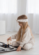 Profile view of a young child wearing a matching oat jogger set from Milana Label, sitting on the floor in a brightly lit room while playing with a wooden car toy.
