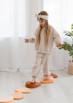A young girl wearing a cozy, oversized oat-colored jogger set and a matching bow headband carefully steps along a row of wooden balancing blocks in a bright room with white sheer curtains.