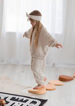 A young girl wearing a beige oversized crewneck with matching joggers while she walks along stepping stones.