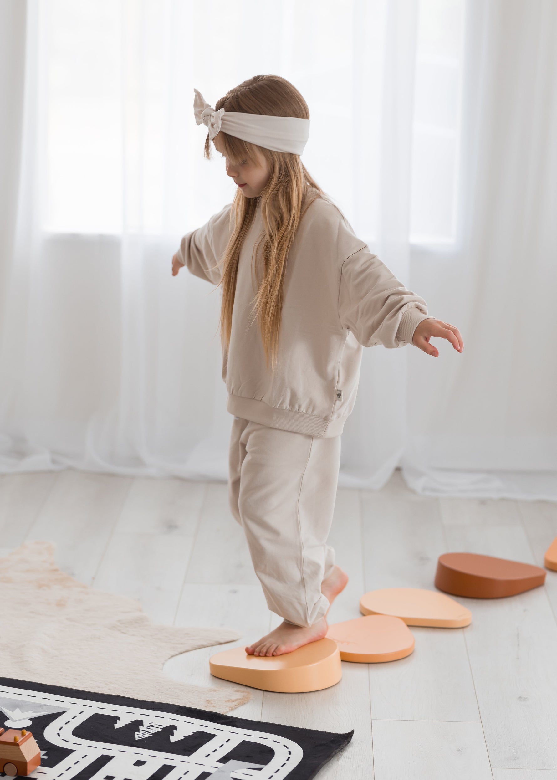 A young girl wearing a beige oversized crewneck with matching joggers while she walks along stepping stones.
