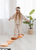 A young girl wearing a cozy oat beige two-piece sweater and jogger set steps across stepping stone blocks in a bright, neutral-toned kids' playroom.