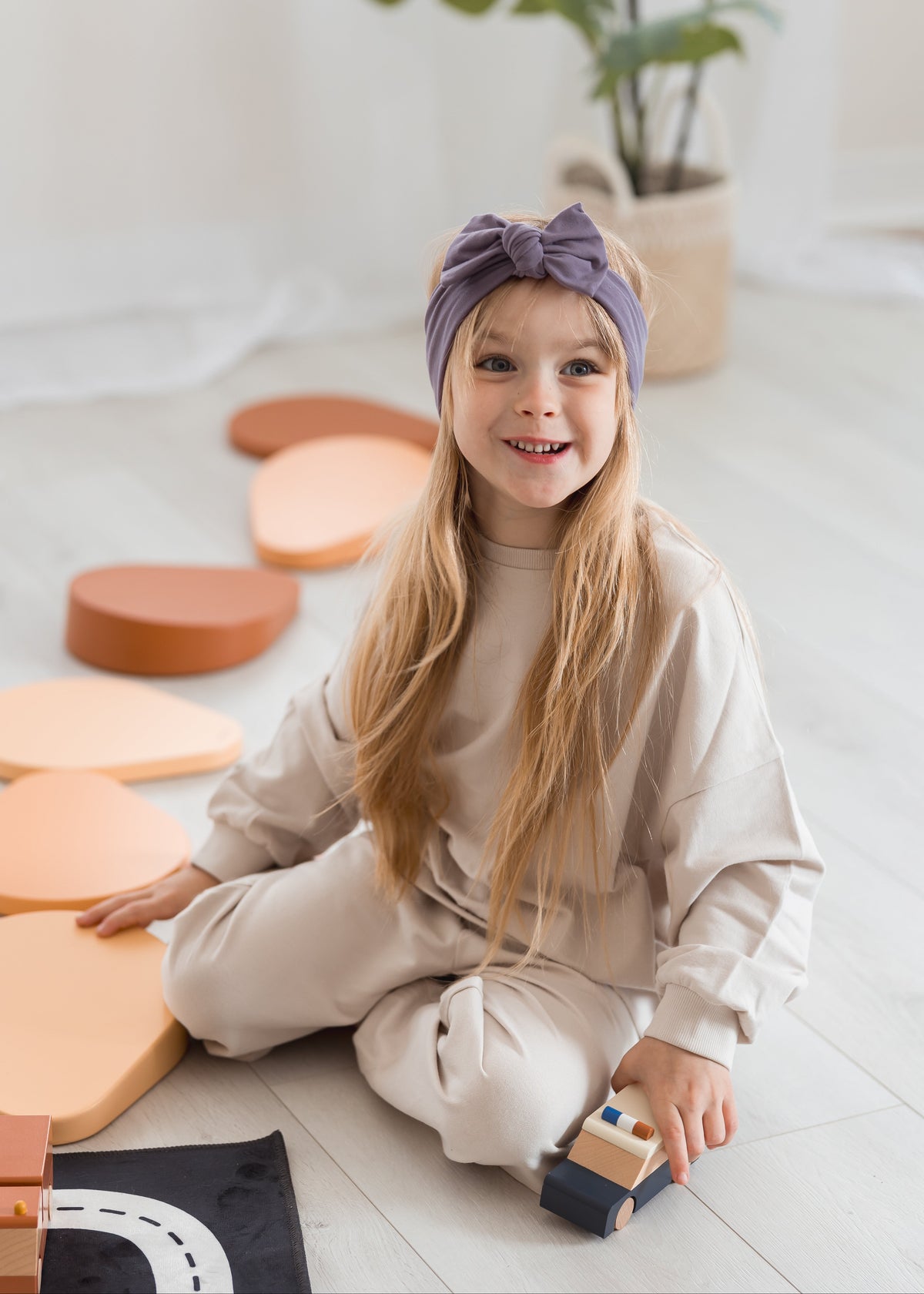 A young girl with long blonde hair sits on a light-colored wooden floor, wearing a beige sweatshirt and matching pants with a purple bow headband. She is holding a small wooden toy car, with pastel-colored geometric blocks scattered around her.