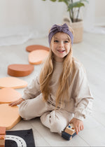 A young girl with long blonde hair sits on a light-colored wooden floor, wearing a beige sweatshirt and matching pants with a purple bow headband. She is holding a small wooden toy car, with pastel-colored geometric blocks scattered around her.