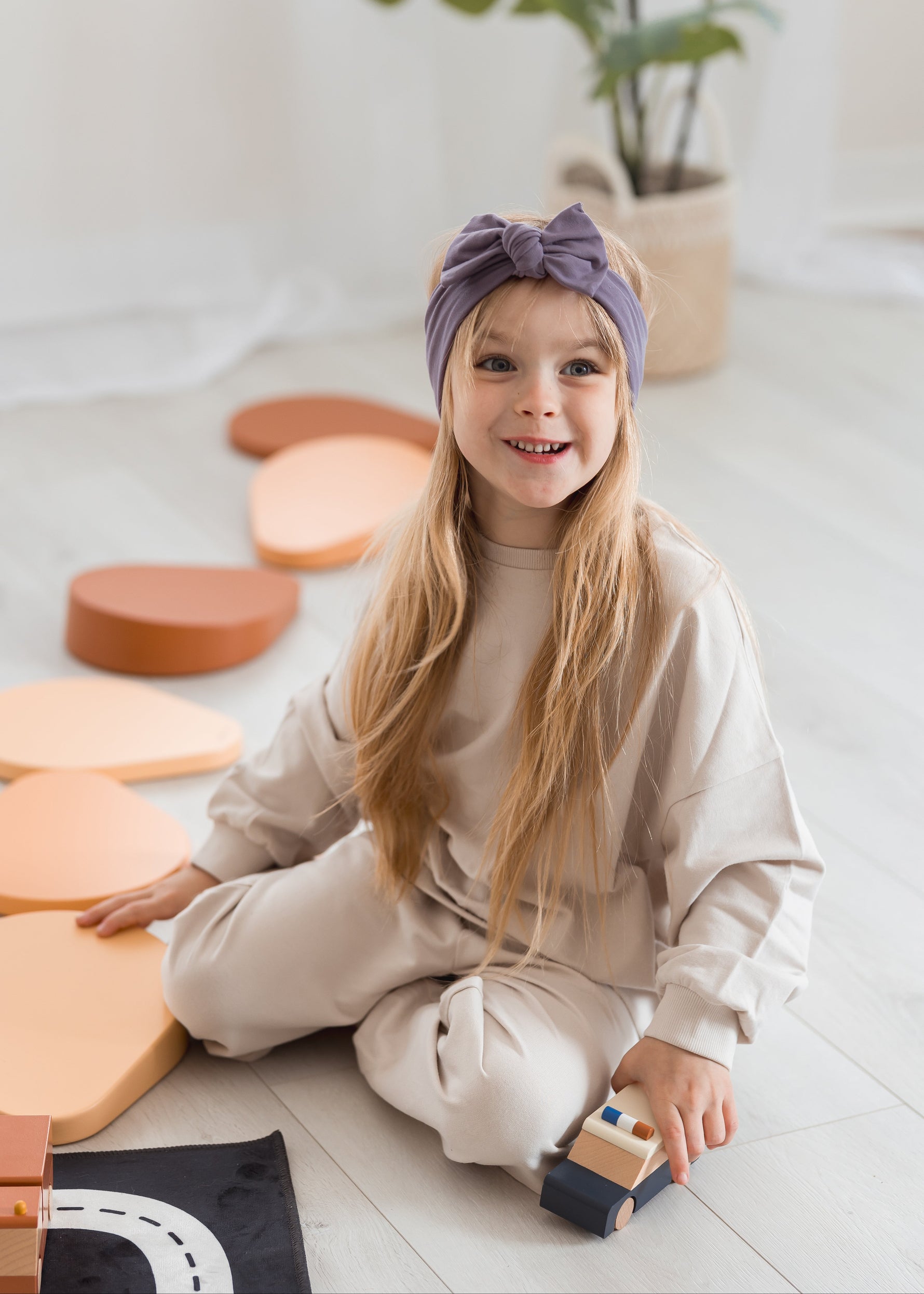 A young girl with long blonde hair sits on a light-colored wooden floor, wearing a beige sweatshirt and matching pants with a purple bow headband. She is holding a small wooden toy car, with pastel-colored geometric blocks scattered around her.