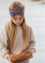 A kid playing with steppers on the floor while modelling the Milana Label knot headband in the colour Wild Mulberry.
