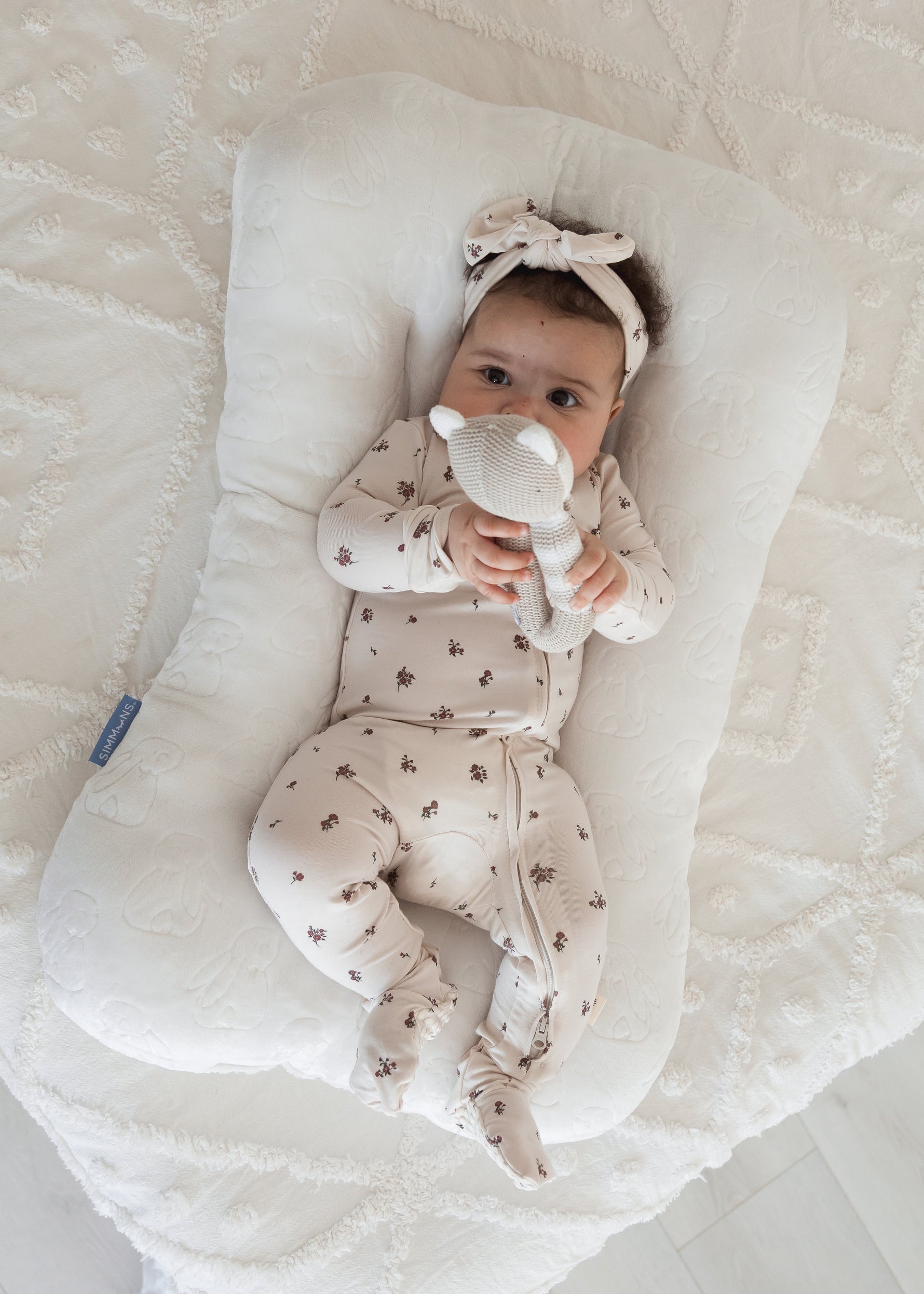 A baby girl playing with a rattle while lounging in her Milana TENCEL sleeper and matching headband in the print Birch Floral.