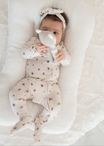 A baby lying on a white nursing pillow, wearing a beige-colored footed sleeper with a small floral pattern, a matching bow headband, and holding a white plush toy.