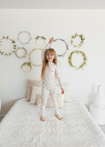 A young girl with long blonde hair stands joyfully on a white bed with her arms raised, wearing a two-piece Tencel PJ set in a delicate birch floral print. The white wall behind her is decorated with minimalist green leaf wreaths.