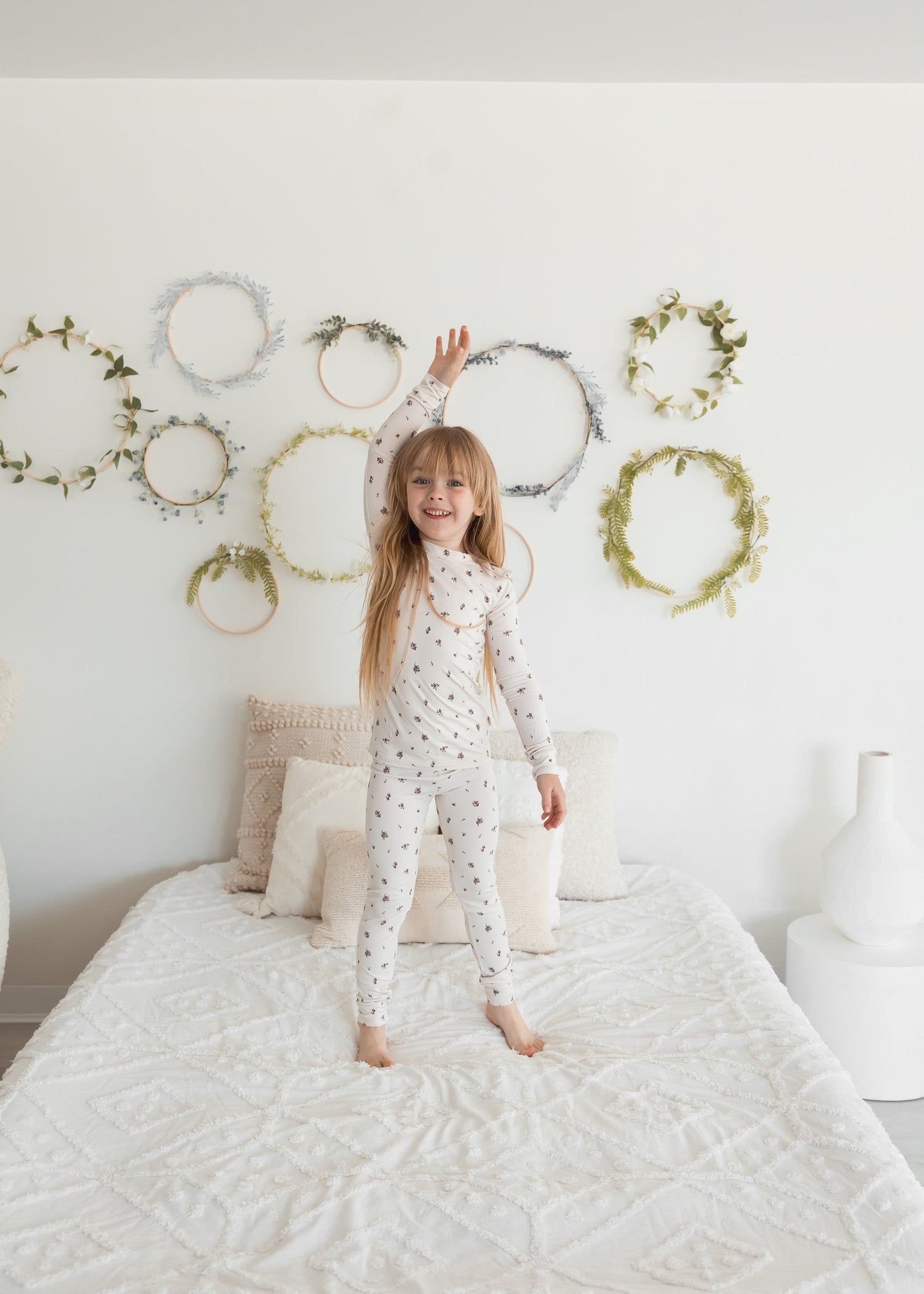 A young girl with long blonde hair stands joyfully on a white bed with her arms raised, wearing a two-piece Tencel PJ set in a delicate birch floral print. The white wall behind her is decorated with minimalist green leaf wreaths.