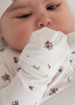 A close-up of a baby wearing a long-sleeve footie and matching hat, both decorated with a small floral pattern, lying on a soft white pillow.