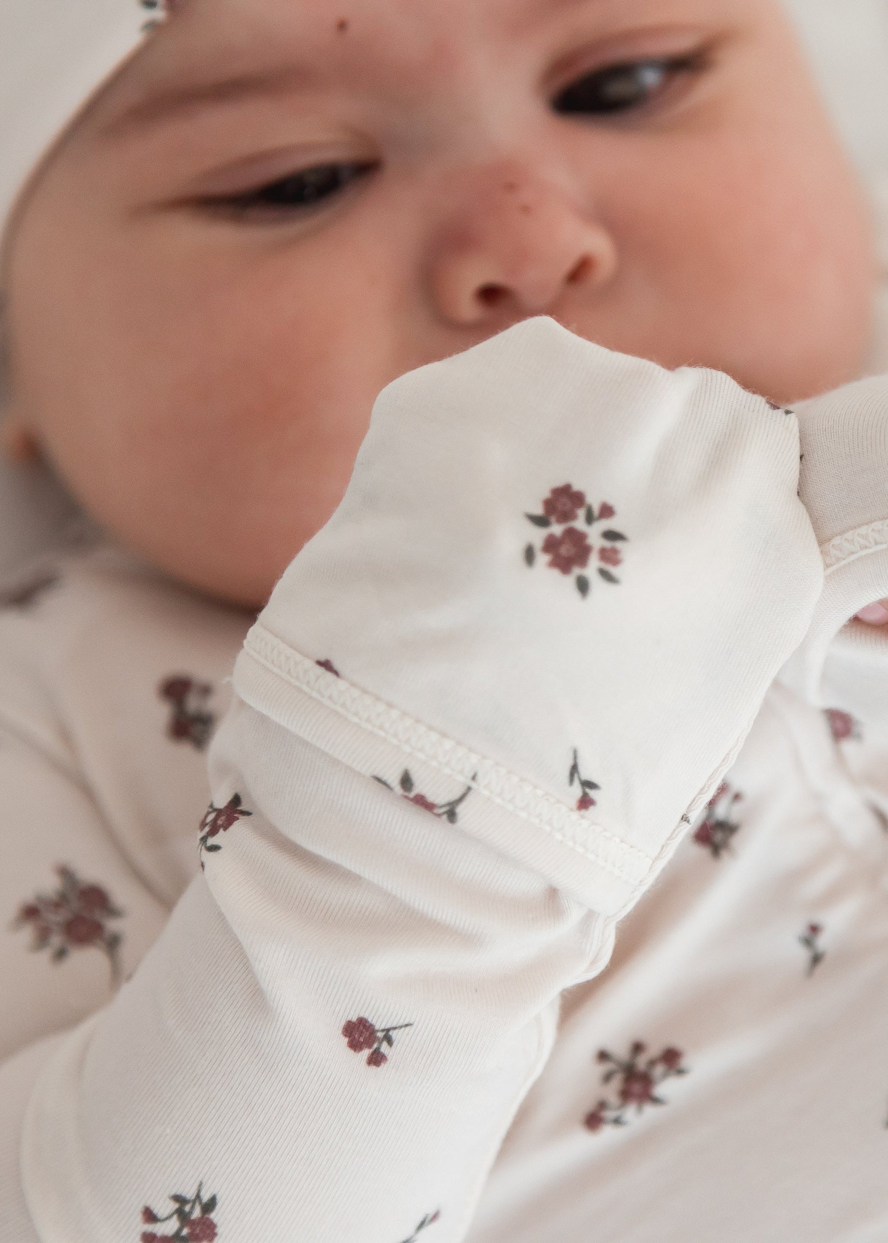 A close-up of a baby wearing a long-sleeve footie and matching hat, both decorated with a small floral pattern, lying on a soft white pillow.