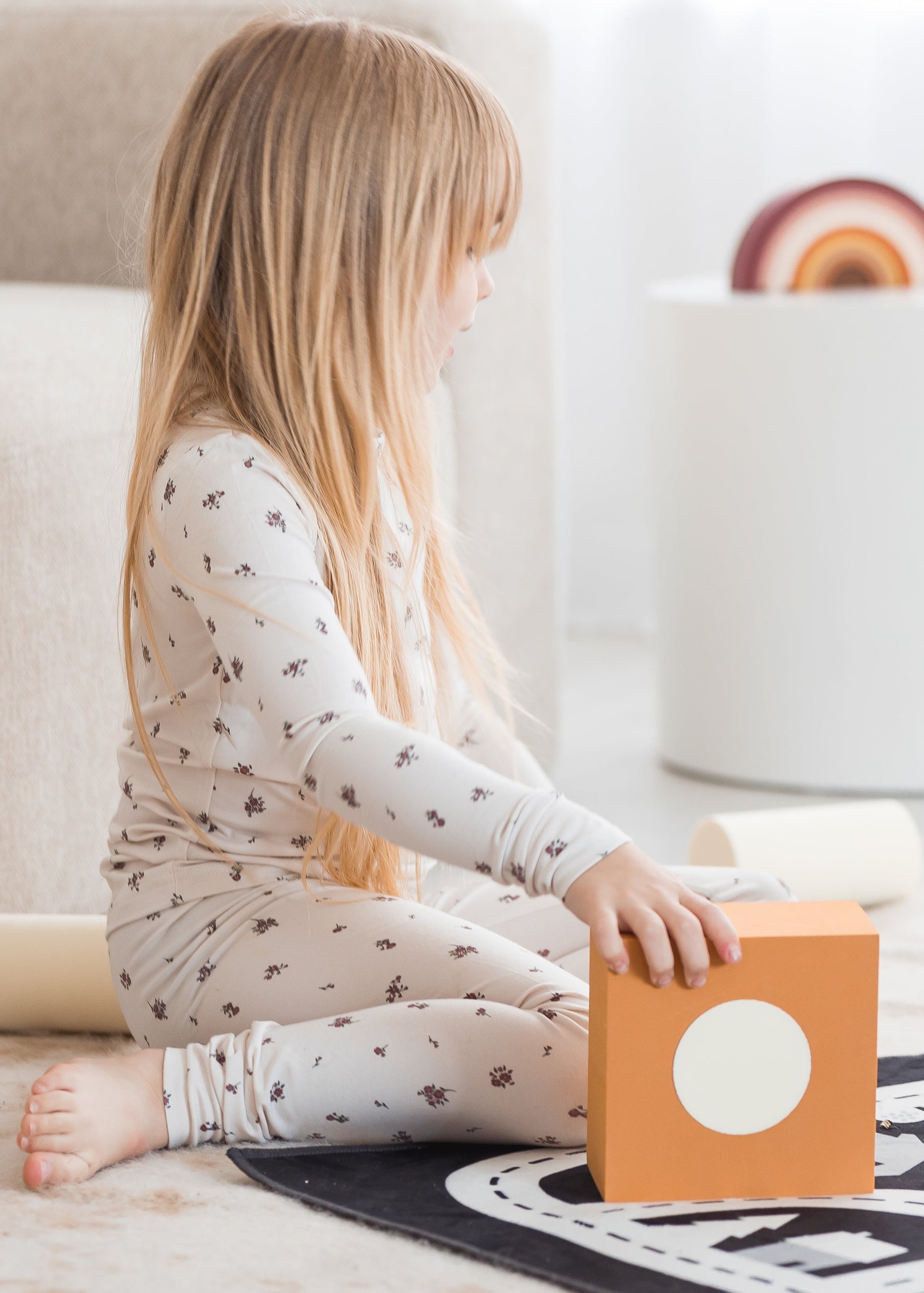 A young girl in a birch floral PJ set sitting on a patterned play mat, playing with neutral-toned foam building blocks in a bright room.