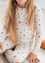 A child wearing a neutral colored long-sleeve pajama set with a small floral pattern, sitting on a light-colored wooden floor.