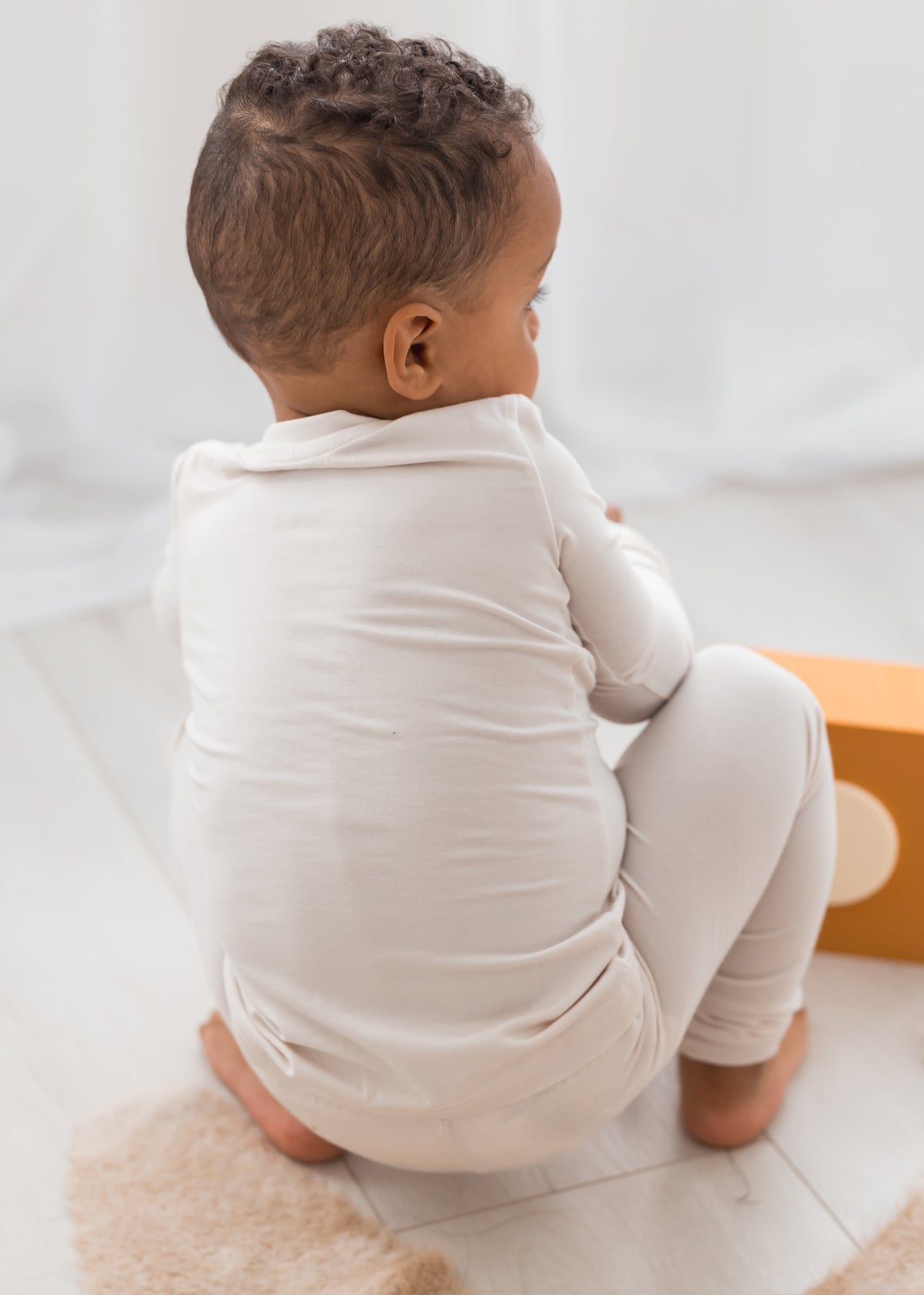 A toddler wearing a light-colored long-sleeve PJ set, sitting on a fluffy beige rug in a bright indoor setting.