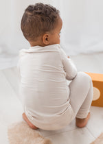 A toddler wearing a light-colored long-sleeve PJ set, sitting on a fluffy beige rug in a bright indoor setting.