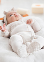 Full-body view of a baby lying in a white lounger wearing a beige zipper baby pajama with grip-dot feet and a matching headband.