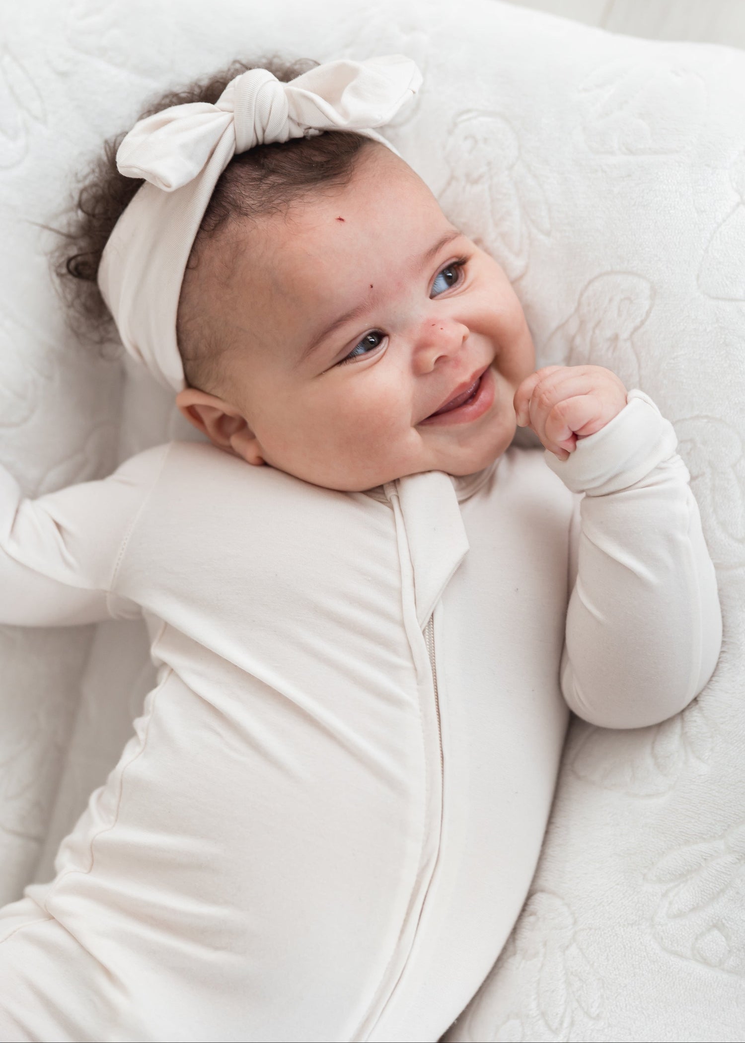 A baby smiling while laying in a neutral lounger wearing a Milana Soft knot headband in the colour Birch.
