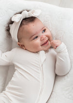 A baby smiling while laying in a neutral lounger wearing a Milana Soft knot headband in the colour Birch.