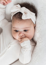 Close-up of a baby wearing a beige TENCEL bow headband and matching sleeper while resting on a white cushion.