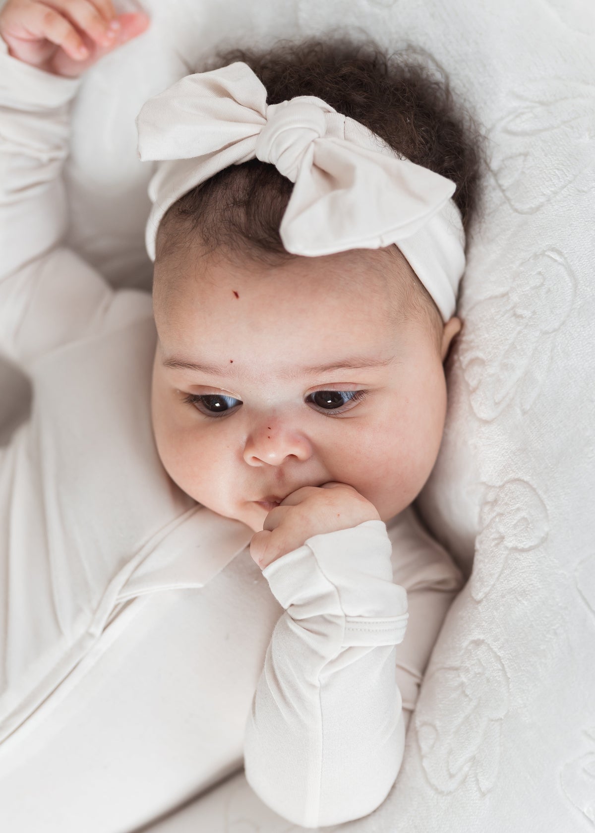 A close-up of a baby wearing a neutral long-sleeve sleeper and a matching bow headband, lying on a textured white blanket.