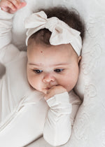A close-up of a baby wearing a neutral long-sleeve sleeper and a matching bow headband, lying on a textured white blanket.