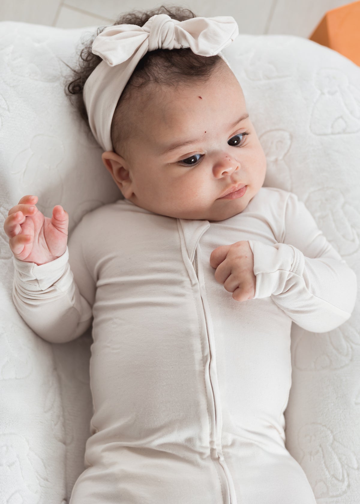 A baby lying on a white quilted cushion, wearing a neutral long-sleeve footie and a matching bow headband.