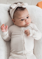 A baby lying on a white quilted cushion, wearing a neutral long-sleeve footie and a matching bow headband.