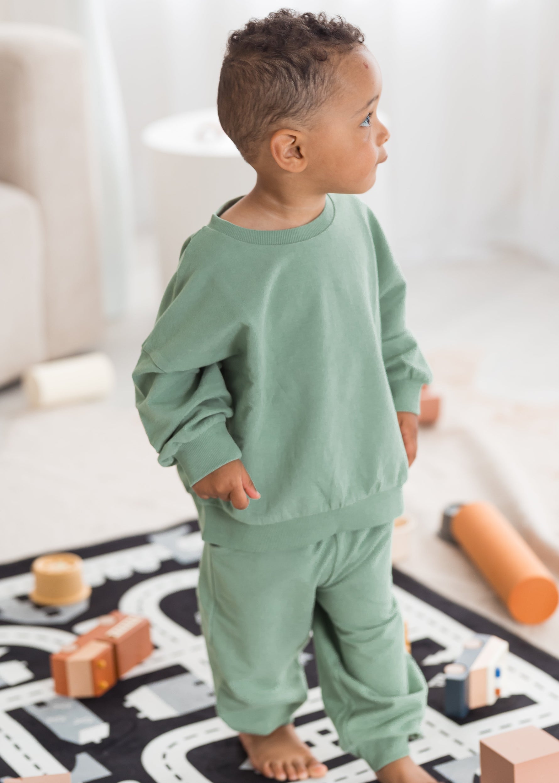 A young toddler boy stands on a road-patterned play mat in a bright, sunlit room, wearing a loose-fit eucalyptus green organic cotton sweatshirt and jogger pants set.