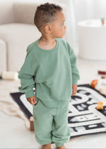 A toddler standing in a bright, modern room wearing a eucalyptus green two-piece lounge set consisting of a long-sleeve sweatshirt and matching sweatpants.