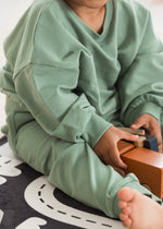 Closeup view of a child sitting on the floor wearing a cozy, oversized eucalyptus green organic cotton tracksuit while playing with wooden blocks on a monochrome play mat.