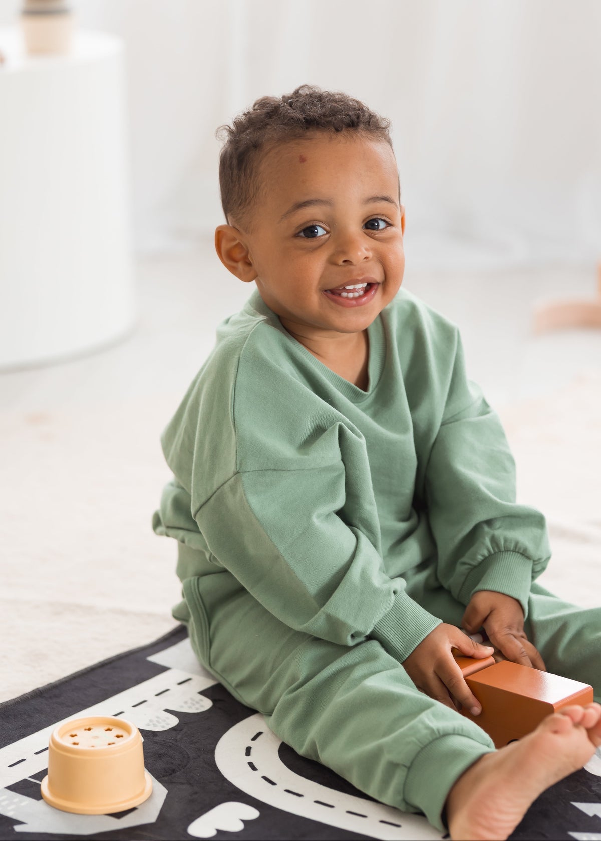 A happy young boy sits on a play mat and smiles toward the camera, wearing a matching muted eucalyptus green organic cotton two-piece jogger set.