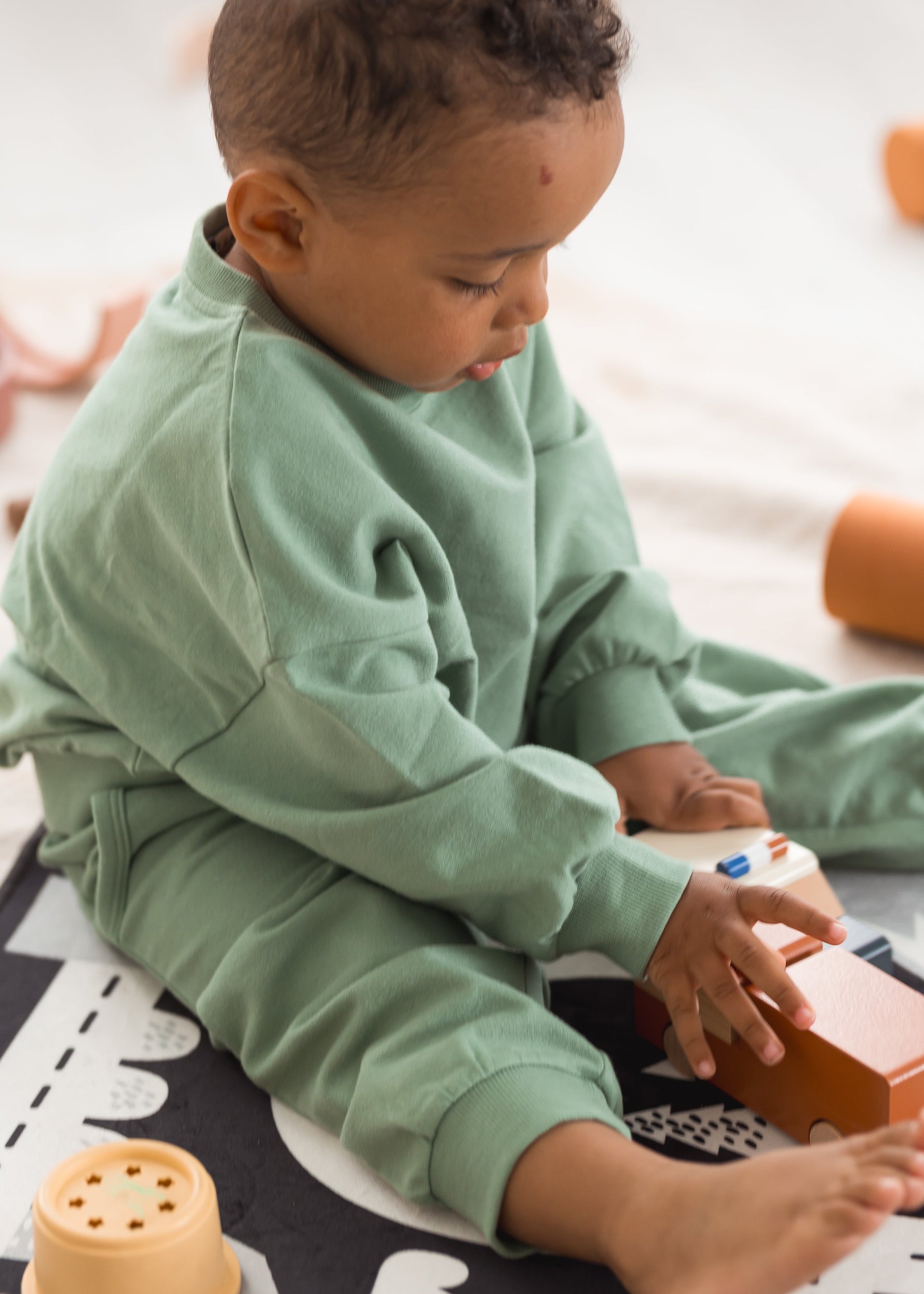 Profile view of a young boy wearing a sage eucalyptus green organic cotton crewneck sweater and pants set, sitting on a play mat and playing with a wooden car.