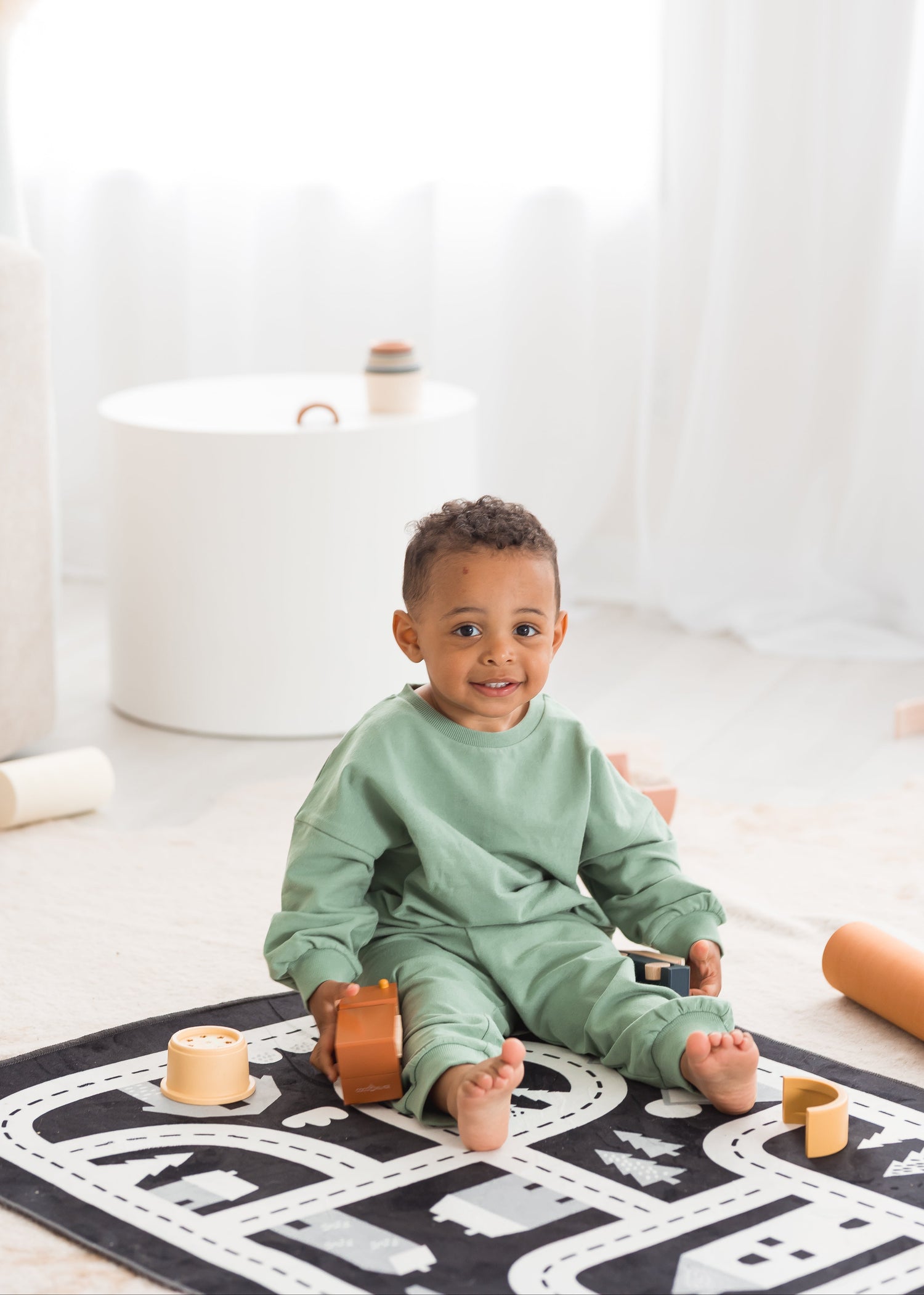 A young toddler boy wearing a matching eucalyptus green organic cotton lounge set sits on a road-patterned play mat in a bright, minimalist room surrounded by wooden toys.