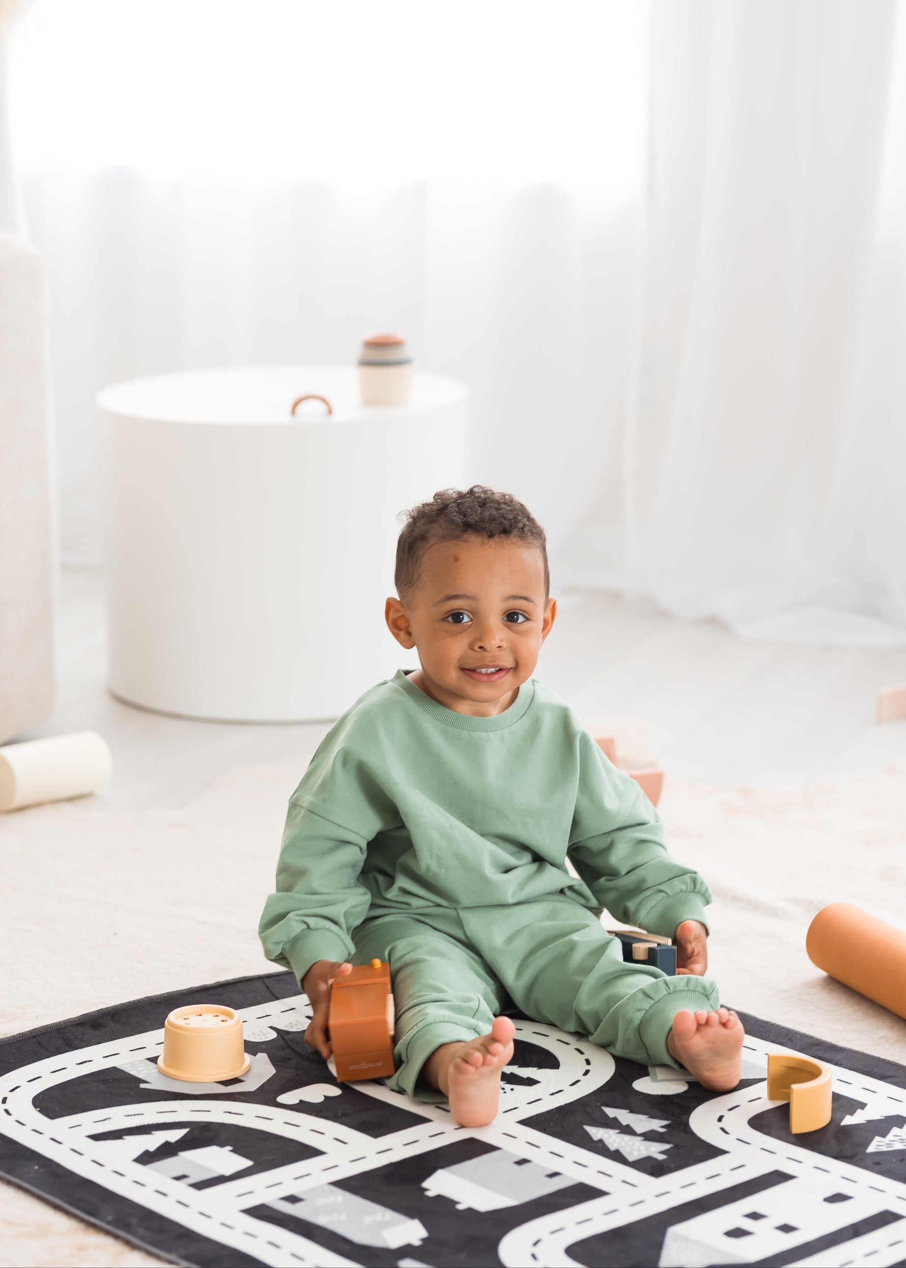 A young toddler boy wearing a matching eucalyptus green organic cotton lounge set sits on a road-patterned play mat in a bright, minimalist room surrounded by wooden toys.