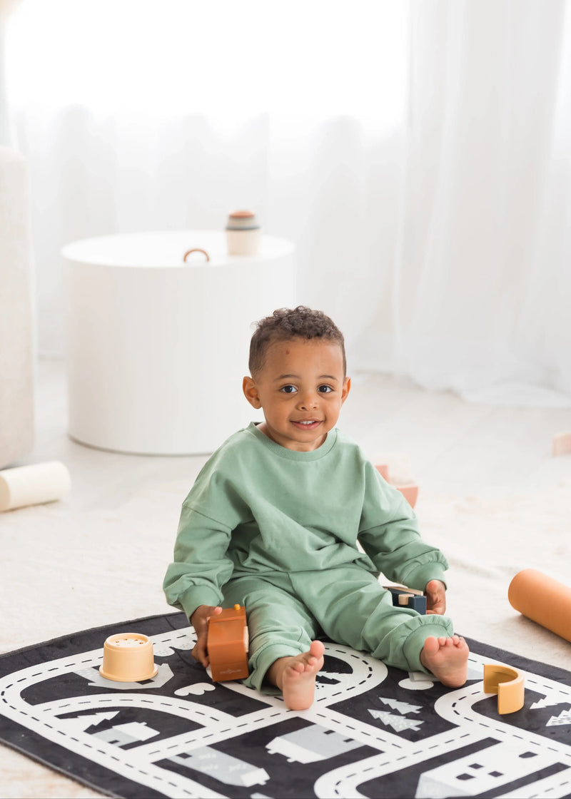 A young toddler boy wearing a matching eucalyptus green organic cotton lounge set sits on a road-patterned play mat in a bright, minimalist room surrounded by wooden toys.
