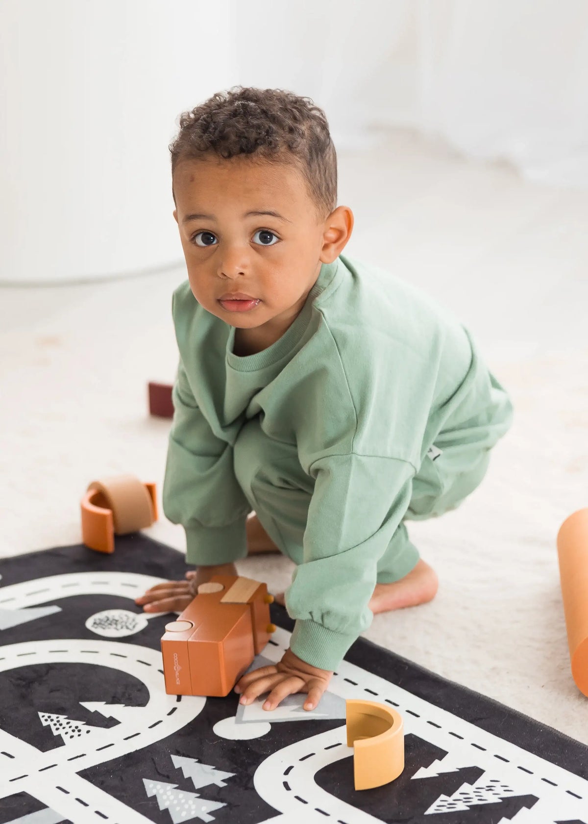 A toddler wearing a eucalyptus green crewneck and matching pants crawls on a black road-map print rug while playing with wooden blocks in a bright, modern room.