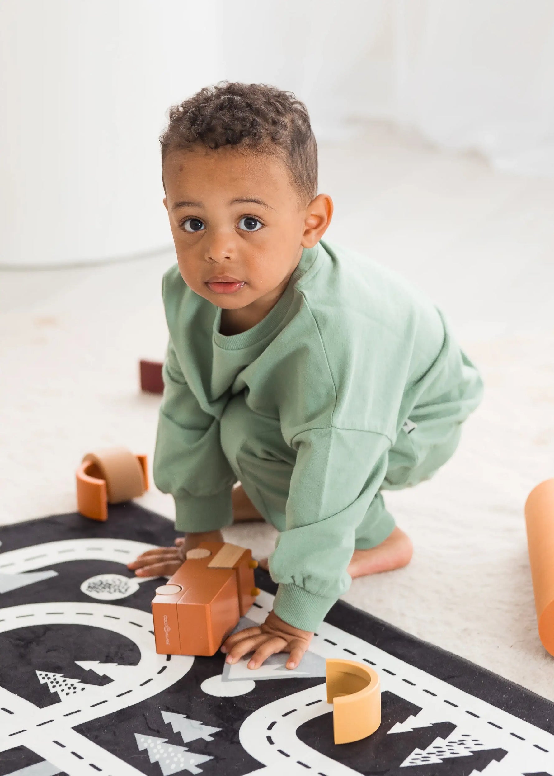 A toddler wearing a eucalyptus green crewneck and matching pants crawls on a black road-map print rug while playing with wooden blocks in a bright, modern room.
