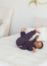 A young boy playfully rolls on a white bed with his feet in the air, wearing a matching dark elderberry purple organic cotton sweatshirt and pajama set.