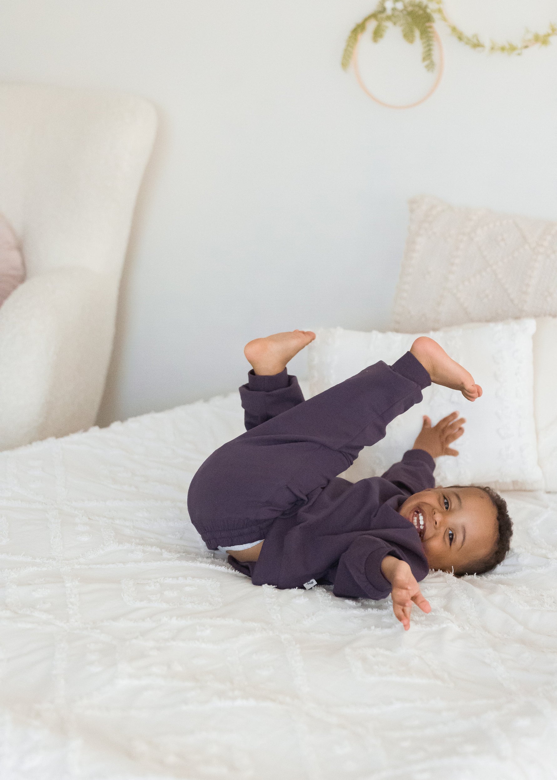 A young boy playfully rolls on a white bed with his feet in the air, wearing a matching dark elderberry purple organic cotton sweatshirt and pajama set.
