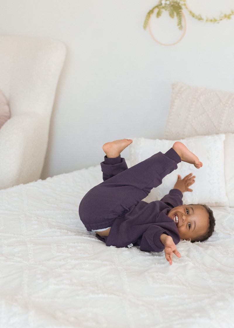 A young boy playfully rolls on a white bed with his feet in the air, wearing a matching dark elderberry purple organic cotton sweatshirt and pajama set.