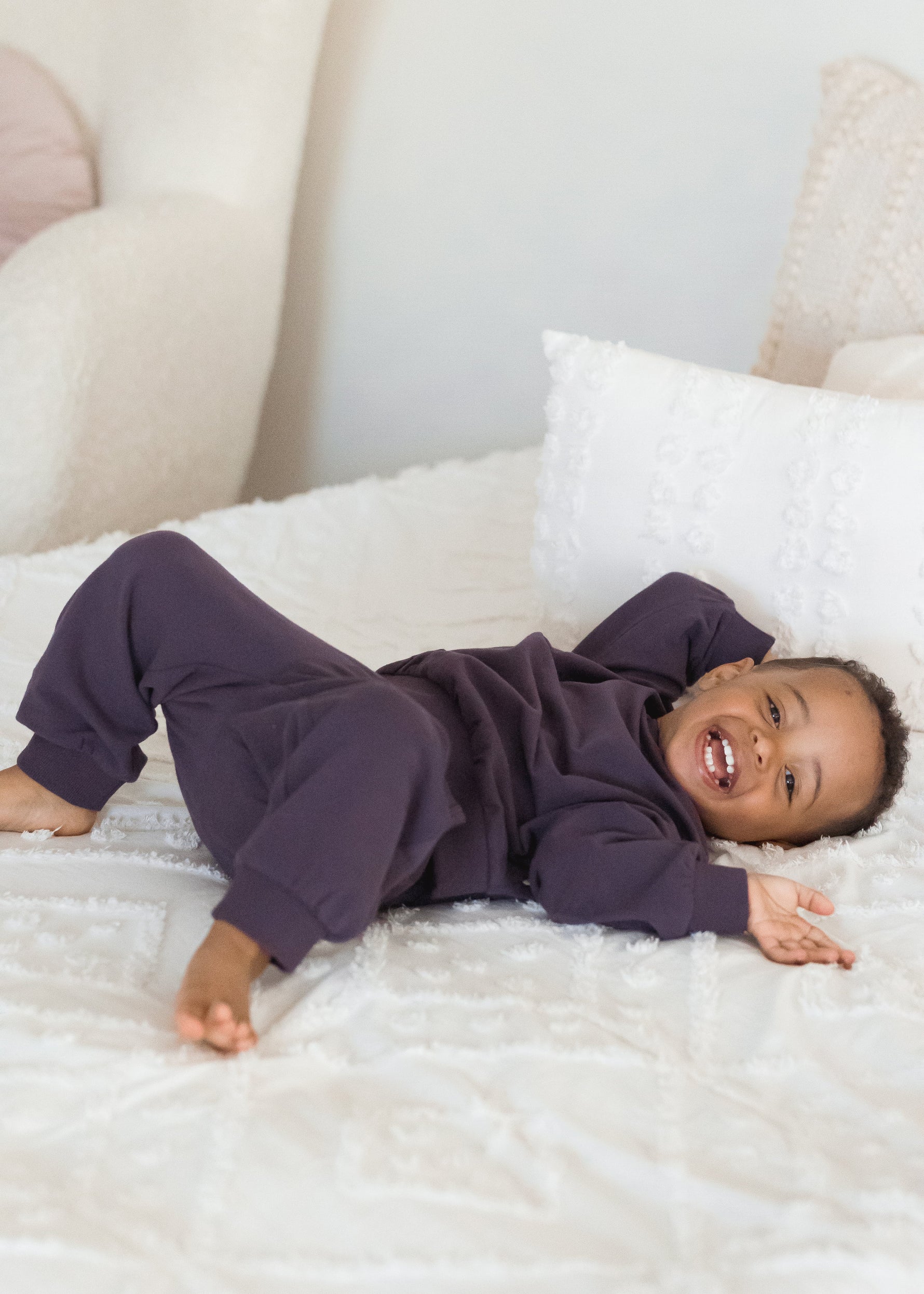 A happy toddler boy lies on his back on a fluffy white bed, smiling widely while wearing a cozy, deep elderberry purple organic cotton two-piece jogger set.