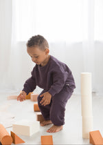 A young boy wearing an elderberry purple organic cotton crewneck sweater and pants lounge set crouches down on a white mat while playing with wooden building blocks.