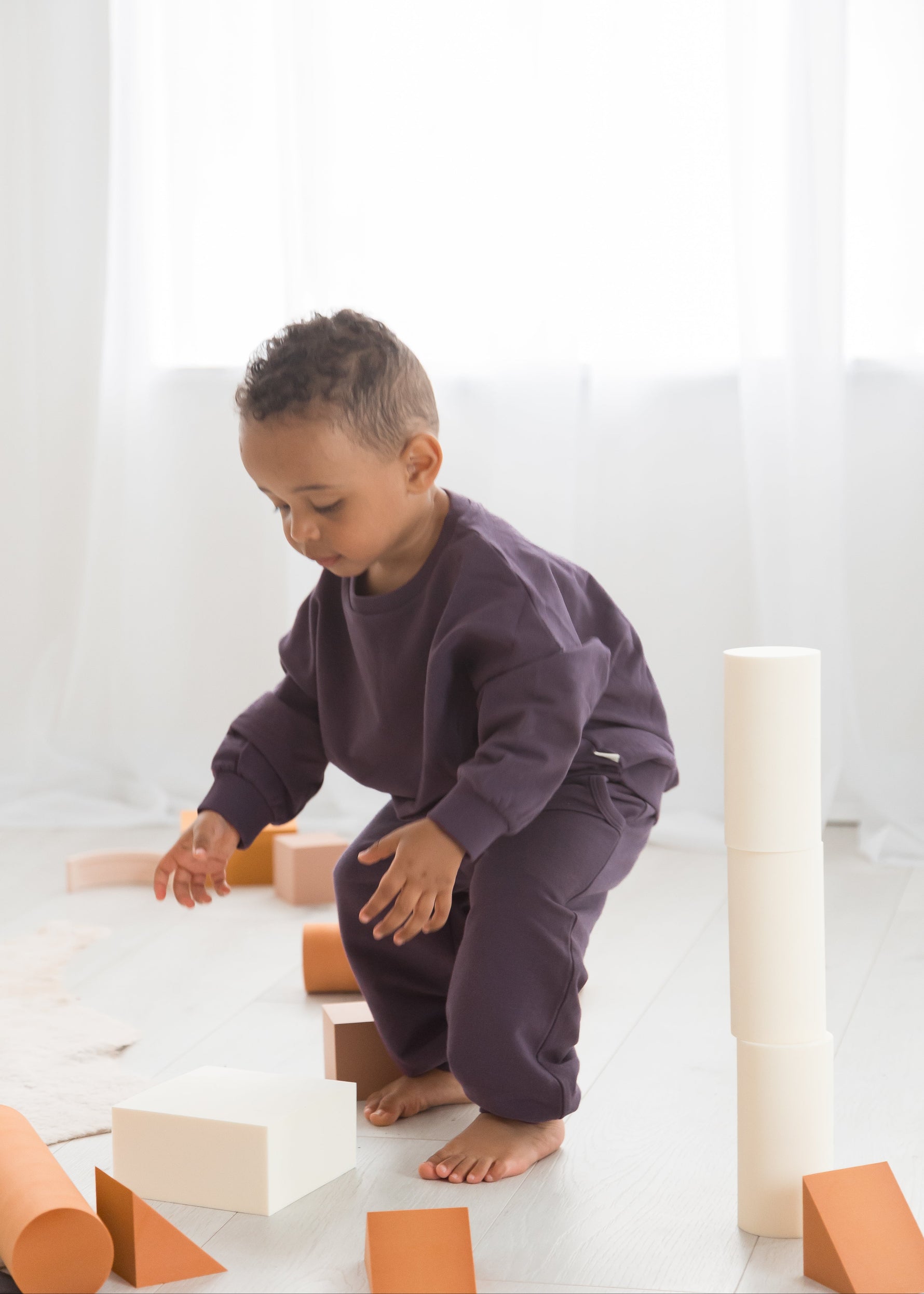 A young boy wearing an elderberry purple organic cotton crewneck sweater and pants lounge set crouches down on a white mat while playing with wooden building blocks.