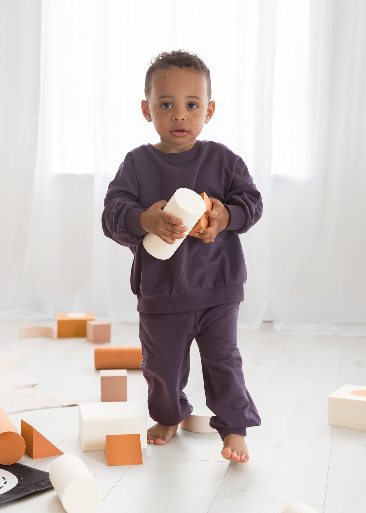 A young boy standing in a brightly lit room wearing a matching deep plum elderberry organic cotton jogger set, holding a white cylindrical wooden toy block.