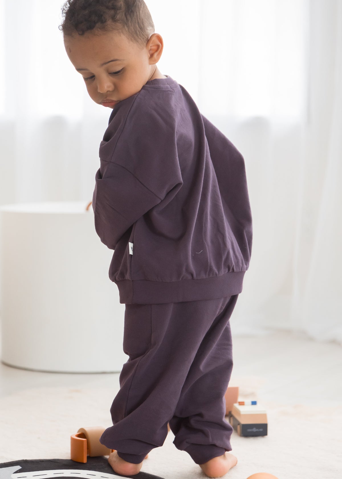 A young toddler boy from the back, wearing a deep elderberry purple organic cotton sweat suit set, standing in a bright minimalist playroom next to wooden toys.