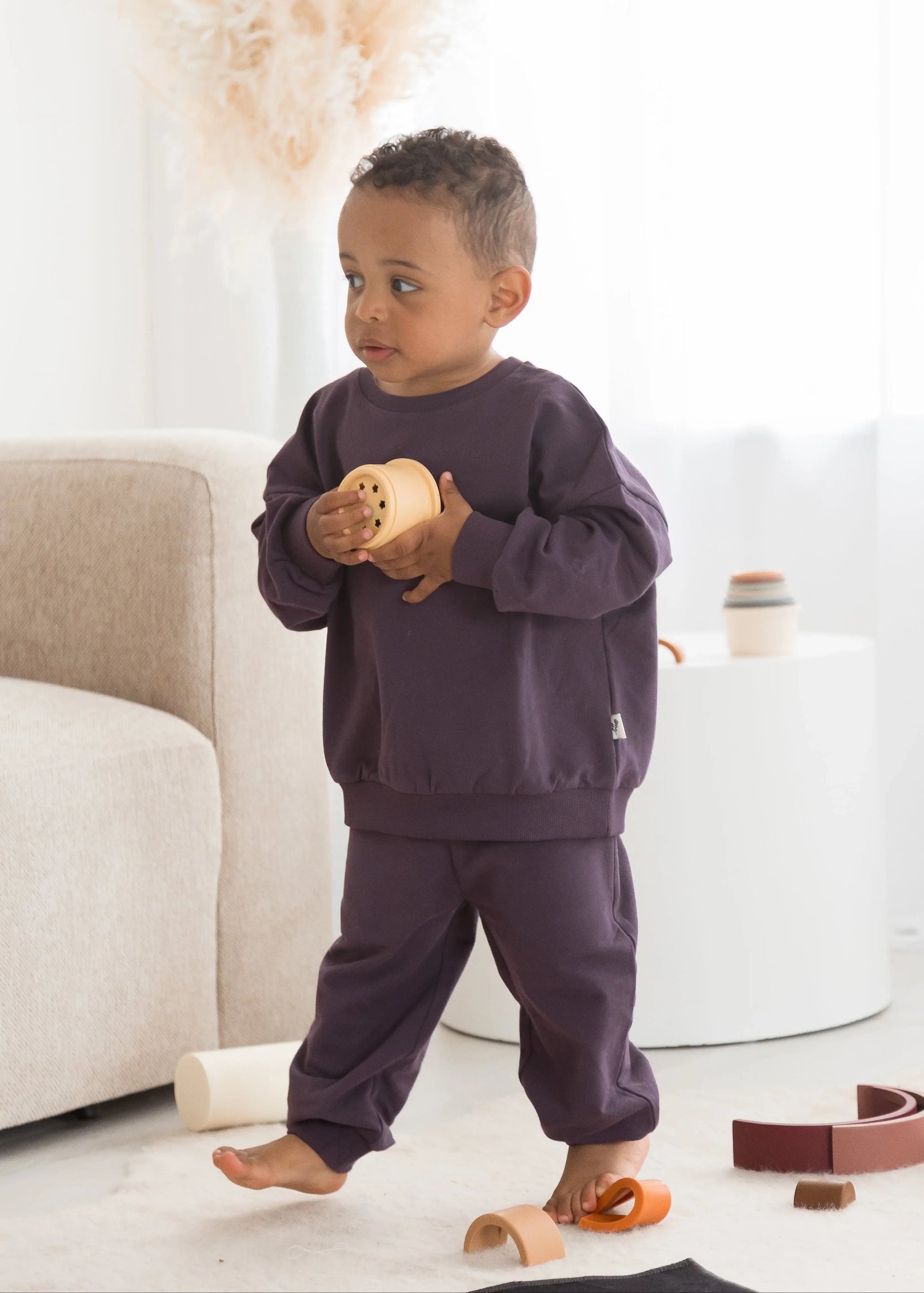 A toddler stands barefoot in a bright, modern room wearing a matching dark purple sweatshirt and sweatpants set, holding a small wooden toy.